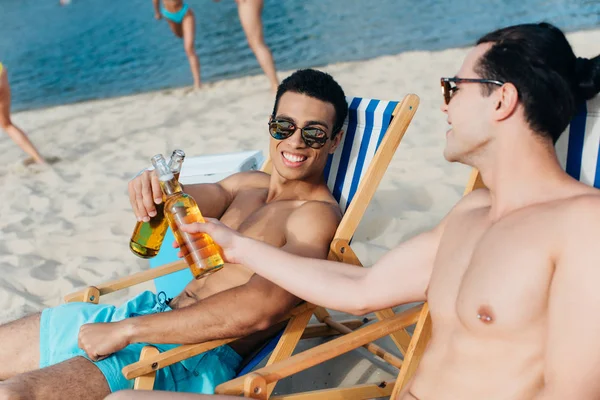 Amigos Multiculturais Alegres Batendo Garrafas Cerveja Enquanto Sentado Salões Chaise — Fotografia de Stock