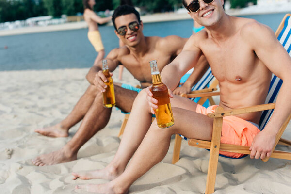 cropped view of young man with mixed race friend smiling at camera while sitting in chaise lounges with bottles of beer