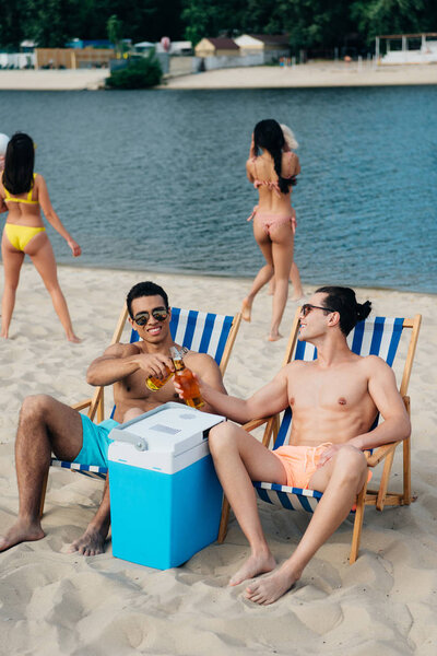 handsome multicultural friends clinking bottles of beer while sitting in chaise lounges near cooler box on beach