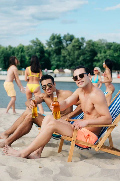 Handsome Multicultural Friends Holding Bottles Beer Smiling Camera While Sitting — Stock Photo, Image