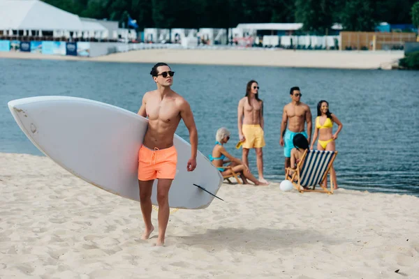 Handsome man holding surfboard while multicultural friends resting on riverside — Stock Image Handsome Man Holding Surfboard While Multicultural Friends Resting Riverside Stock Picture