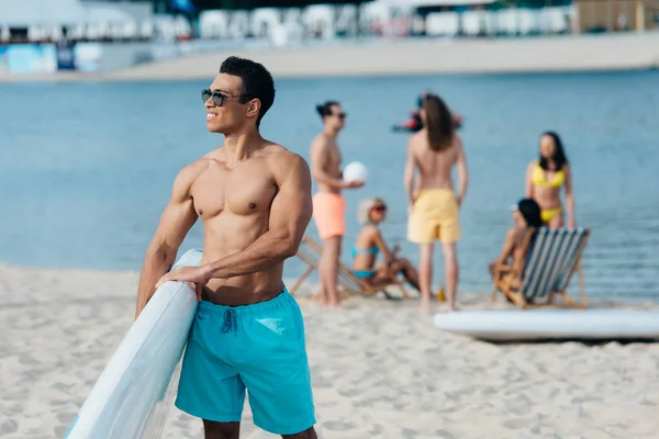 Handsome mixed race man holding surfing board while multicultural friends resting near river on beach — Stock Image Handsome Mixed Race Man Holding Surfing Board While Multicultural Friends Royalty Free Stock Images