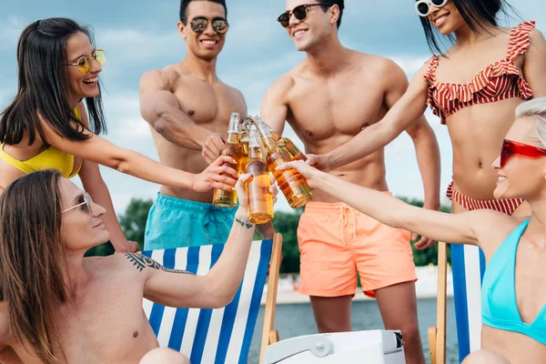 Cheerful multicultural friends clinking bottles of beer while resting on beach — Stock Image Cheerful Multicultural Friends Clinking Bottles Beer While Resting Beach Stock Photo