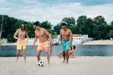 yakışıklı çok kültürlü erkek genç kadınların yakınında plajda futbol oynuyor