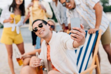 selective focus of cheerful young man taking selfie with multicultural friends having fun on beach clipart