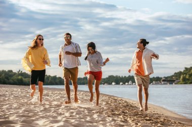 cheerful multicultural friends running on sand beach near river clipart