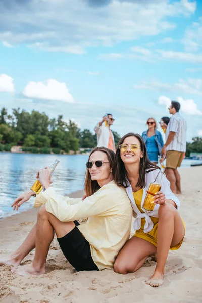 Alegre Jovem Casal Sentado Areia Segurando Garrafas Cerveja Perto Amigos — Fotografia de Stock