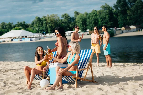Attractive multicultural girls clinking bottles of beer while resting on beach near young men — Stock Image Attractive Multicultural Girls Clinking Bottles Beer While Resting Beach Young Royalty Free Stock Images