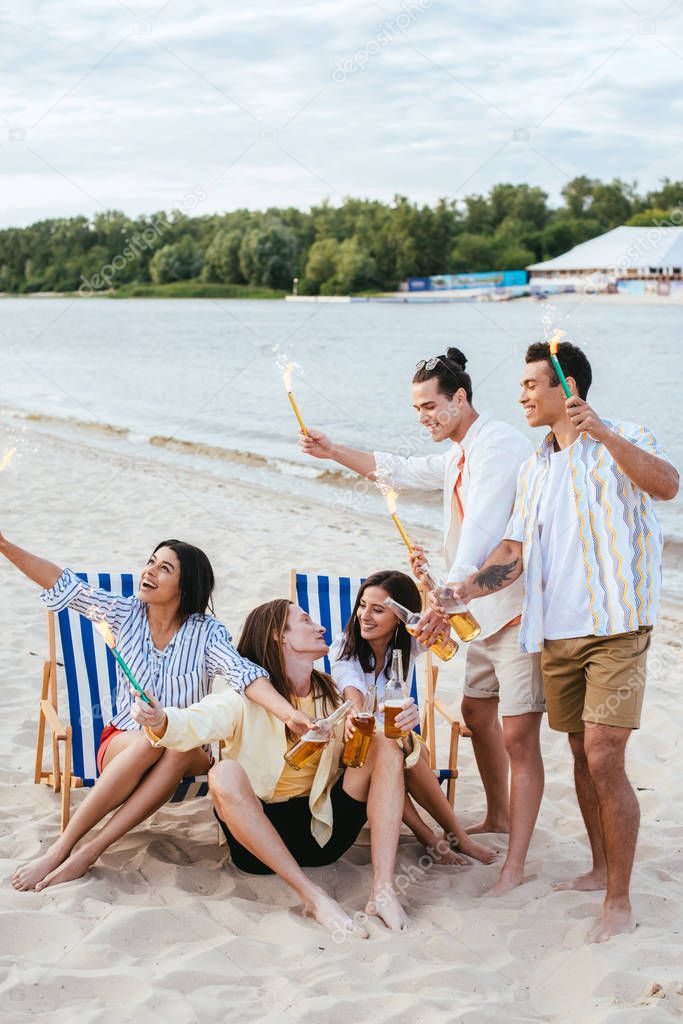 cheerful multicultural friends holding bottles of beer and sparklers while having fun on beach