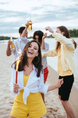 çok kültürlü arkadaşlarının yanında sahilde eğlenirken kıvılcım tutan ve kameraya bakan mutlu genç kadın