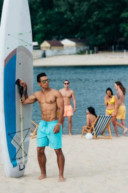 handsome mixed race man looking at camera while standing near surfboard on beach clipart