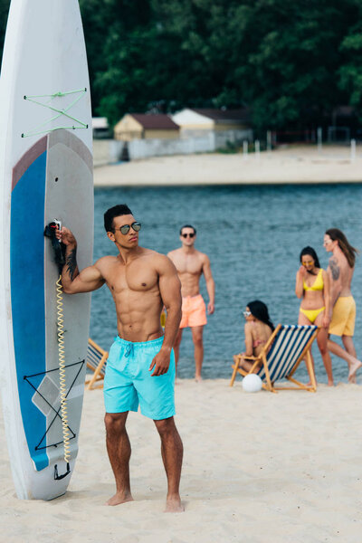 handsome mixed race man looking at camera while standing near surfboard on beach
