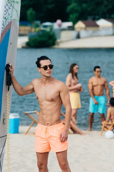 ビーチでサーフボードの近くに立っている間 遠くを見てサングラのハンサムな若者 — ストック写真