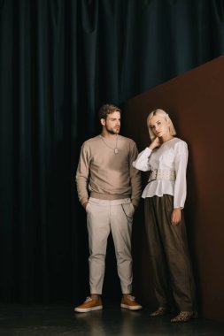 full length view of stylish man with hands in pockets and blonde woman in blouse standing near curtain clipart