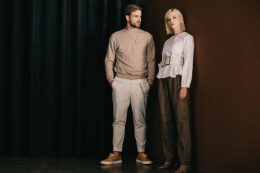 full length view of stylish man with hands in pockets and blonde woman in blouse standing near curtain clipart