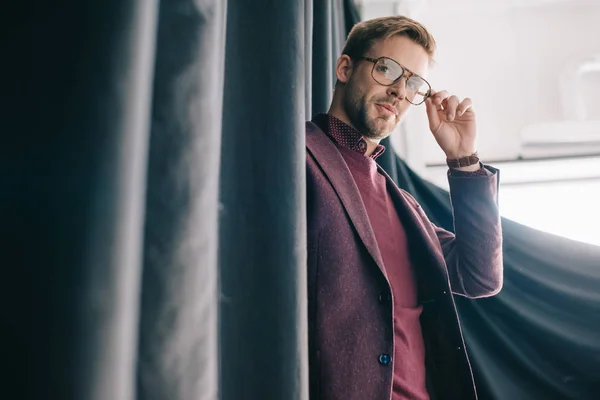 Stylish Young Man Jacket Touching Glasses While Standing Curtain — Stock Photo, Image