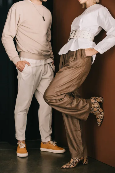 cropped view of stylish man and woman in boots with snakeskin print standing with hands in pockets near curtain