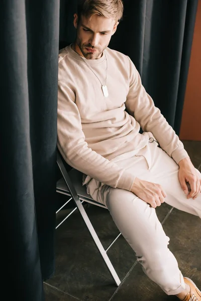 bearded man in casual outfit sitting on chair near curtain