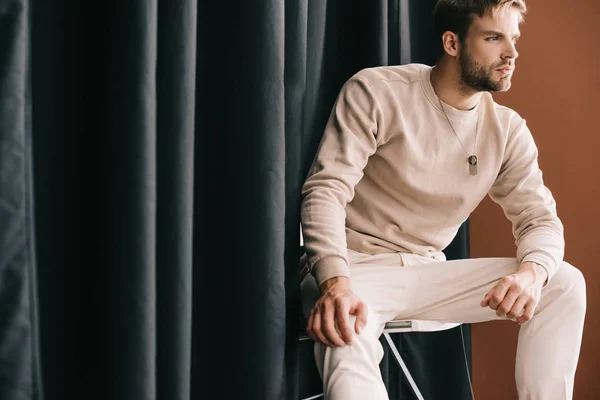 man in casual outfit sitting on chair near curtain on brown