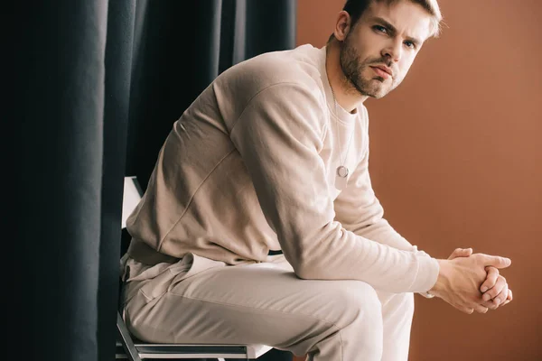 man in casual outfit sitting on chair with clenched hands near curtain on brown