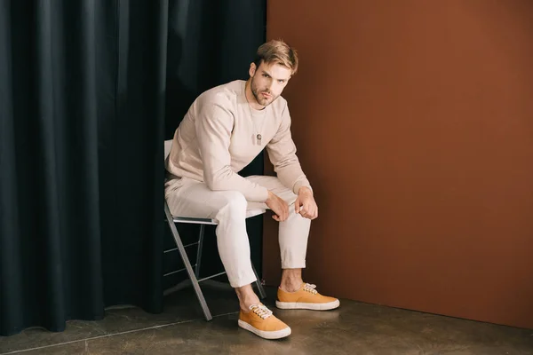 man in casual outfit sitting on chair and looking at camera near curtain on brown