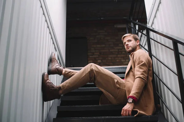 pensive man in brown coat sitting on stairs and looking away