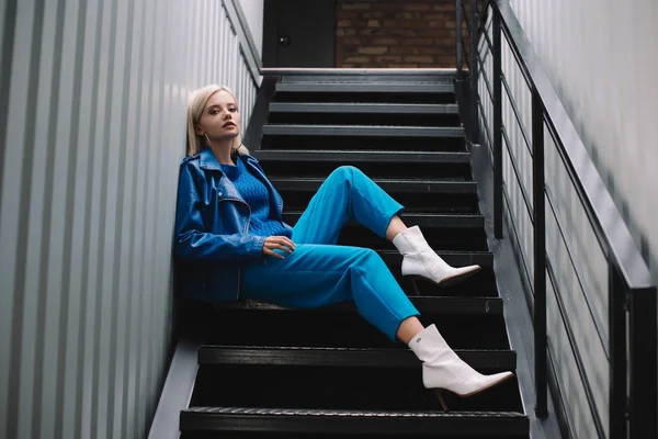 青い革のジャケットと階段に座っているかかとを着ているブロンドの女性 — ストック写真