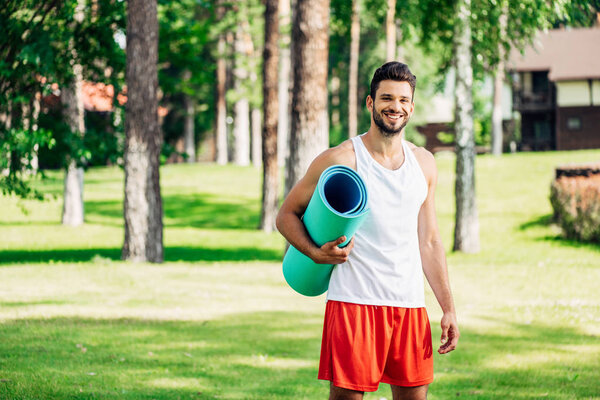 happy athletic sportsman holding fitness mat in park 
