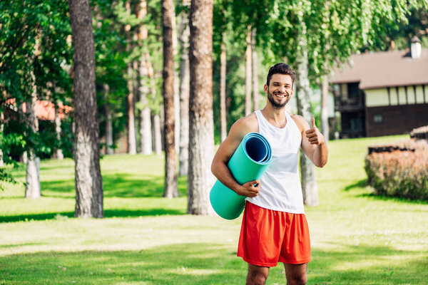 happy athletic sportsman holding fitness mat and showing thumb up 