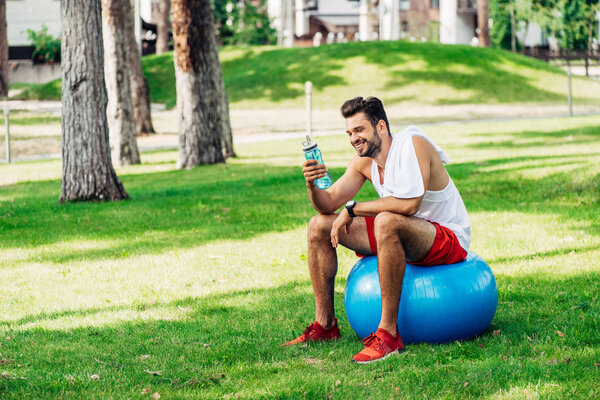 happy bearded man looking at sport bottle while sitting on fitness ball 