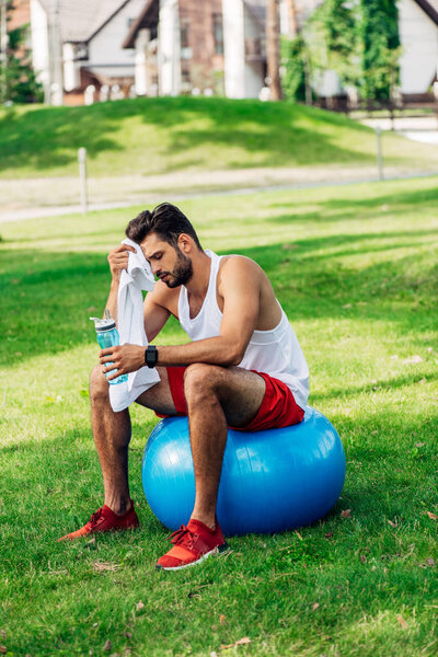 bearded man holding sport bottle while sitting on fitness ball and wiping sweat