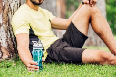ağaç gövdesi yakınında spor şişesi tutan sakallı sporcunun kırpılmış görünümü 
