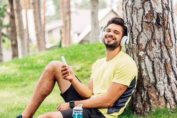 Hombre Feliz Auriculares Sentados Cerca Del Árbol Sosteniendo Teléfono Inteligente — Foto de Stock