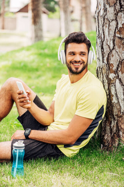positive man in headphones using smartphone near tree trunk and listening music 