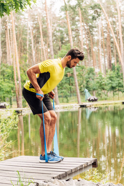 handsome bearded man exercising with suspension straps near lake in forest 