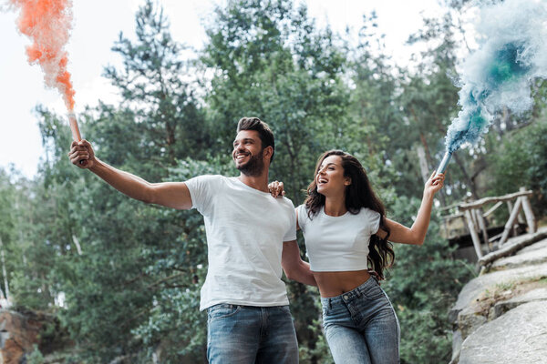 cheerful bearded man and attractive woman holding smoke bombs 