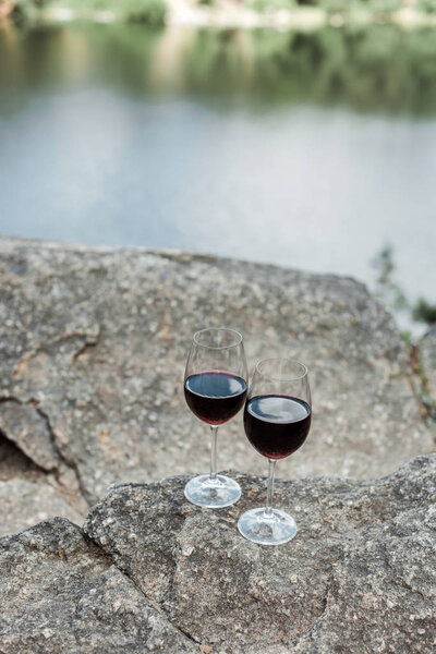 selective focus of wine glasses with red aromatic wine on stone near lake 