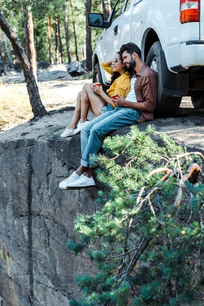 选择性的焦点胡子的男人和有吸引力的女孩坐在车边 在树林里拿着杯子 — 图库照片