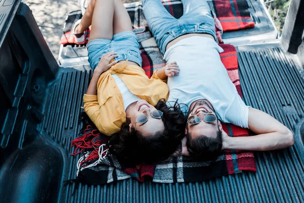 overhead view of woman in sunglasses lying with handsome man in plaid blanket in car trunk