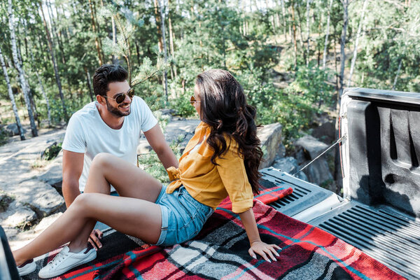 happy man in sunglasses looking at woman while sitting in car trunk in woods 