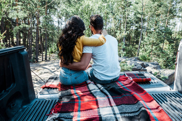 back view of young woman and man hugging while sitting in car trunk in woods 