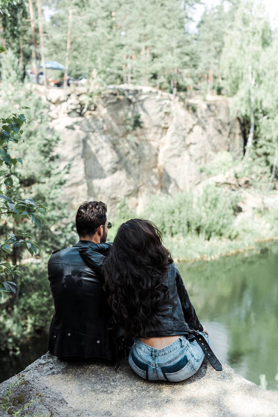 back view of man and woman in leather jackets sitting near trees in woods 