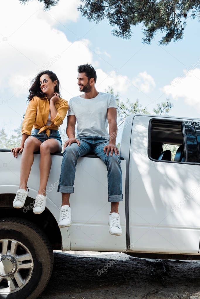 handsome bearded man sitting on car and looking at happy girl 