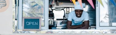 panoramic shot of happy african american man in chef uniform smiling from food truck  clipart