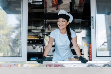 happy asian woman in hat and chef uniform smiling in food truck  clipart