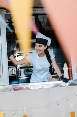 selective focus of cheerful asian woman in hat and chef uniform smiling in food truck  clipart