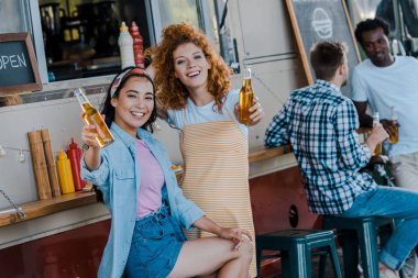 redhead kadın ve gıda kamyonu yakınında bira tutan mutlu asya kız seçici odak