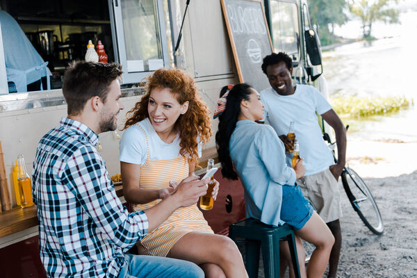 selective focus of handsome man holding smartphone near redhead girl and multicultural people 