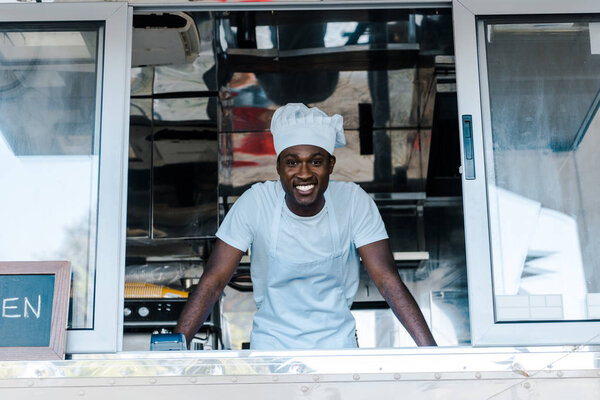 cheerful african american man in chef uniform and hat smiling from food truck 