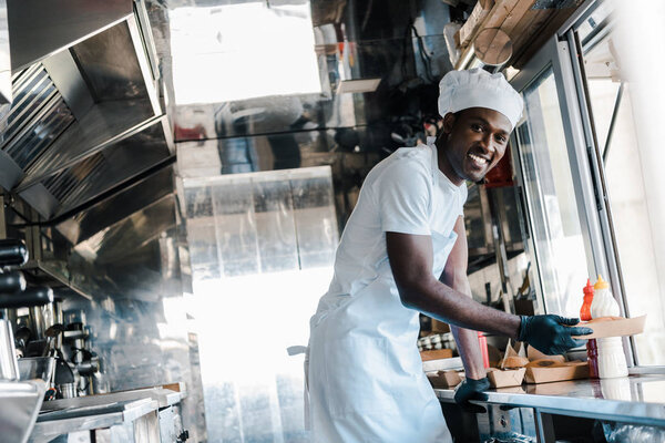 selective focus of cheerful african american chef smiling while holding carton plate in food truck 